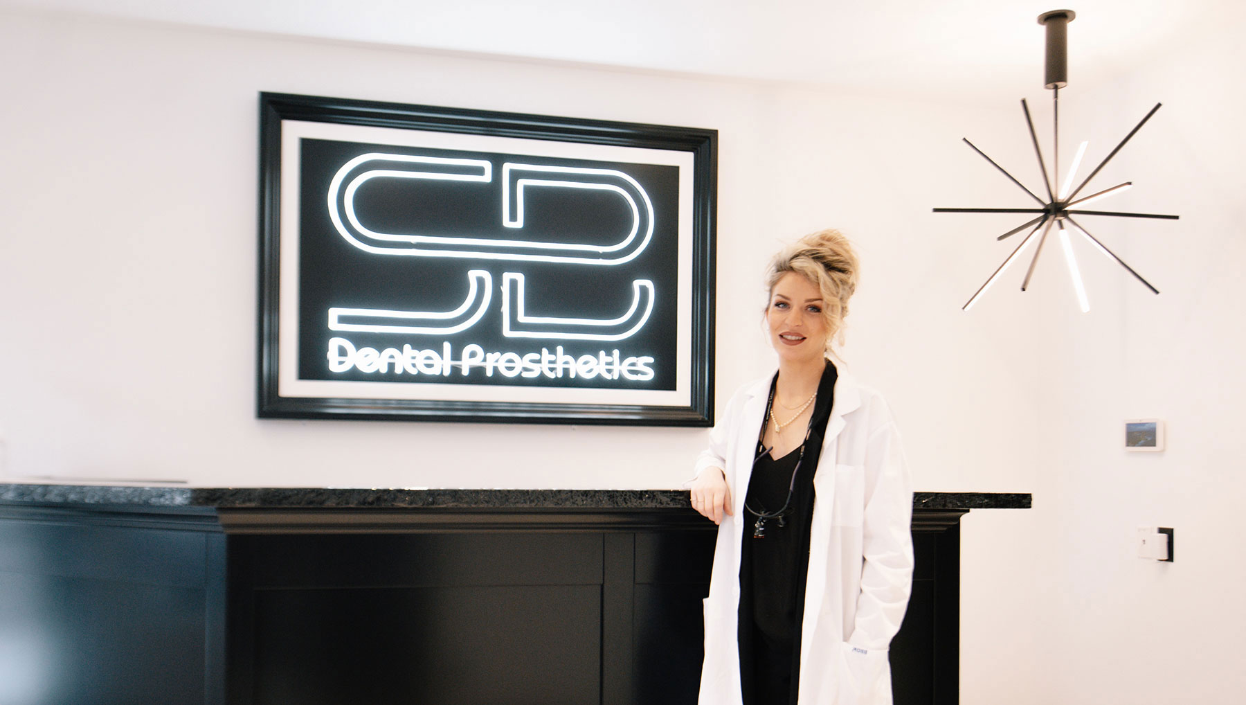 The denturist and owner of SB Denture clinic posing for a photo in front of the reception desk and neon lit company logo close up